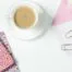 A cup of coffee, a pink notebook, scattered paperclips and an envelope on a white desktop