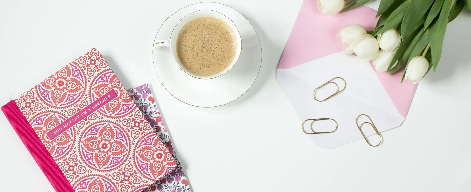 A cup of coffee, a pink notebook, scattered paperclips and an envelope on a white desktop