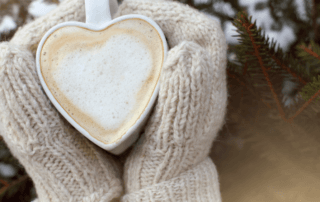 Hands with mittens on warming themselves around a heart shaped mug of frothy coffee or hot chocolate