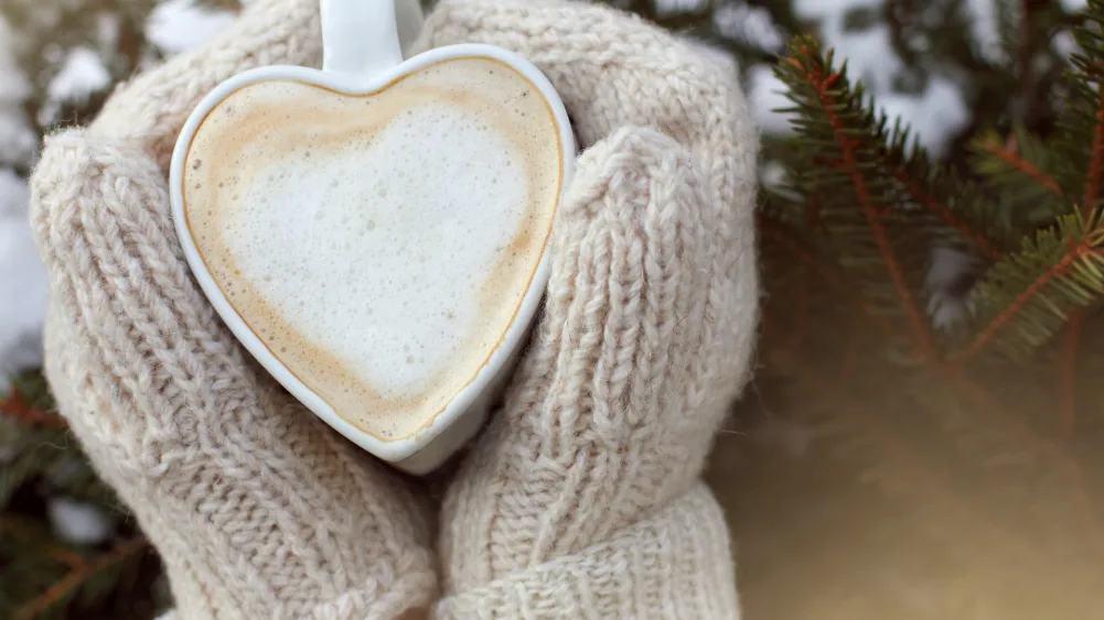 Hands with mittens on warming themselves around a heart shaped mug of frothy coffee or hot chocolate